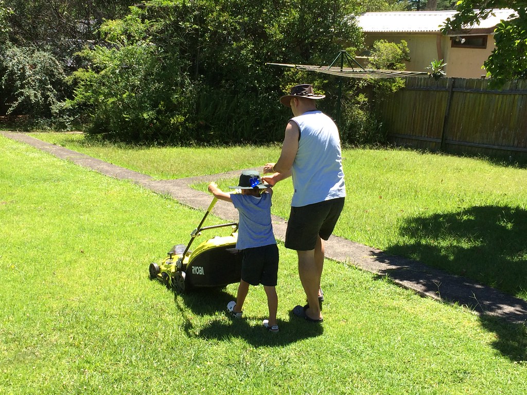 Man and young child pushing a yellow Ryobi mower across a sunny backyard lawn
